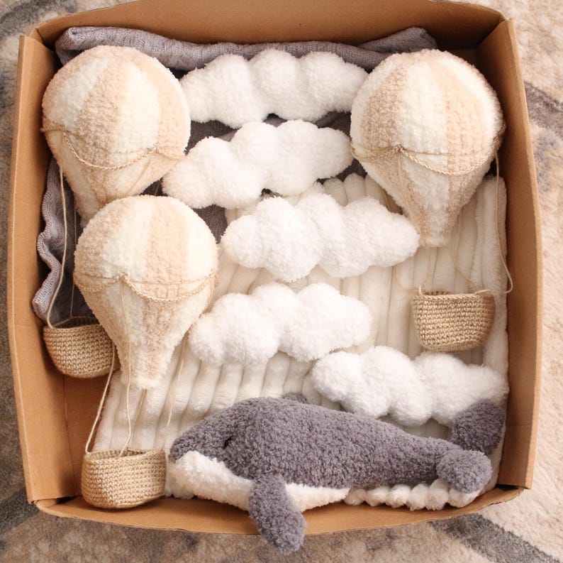 May include: A set of plush toys in a brown box, including three beige hot air balloons, a grey whale, and white fluffy clouds. The balloons have baskets made of woven material.