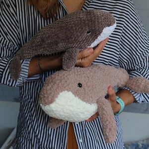 May include: Two crocheted whale plushies, one brown and one light brown, with white accents around the mouth. The whales are being held by a person's hands.