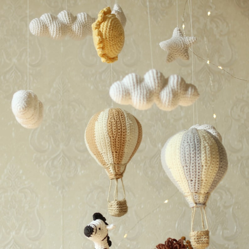 May include: A baby mobile with a crocheted theme. It features a sun, clouds, a star, and two hot air balloons in beige and gray.