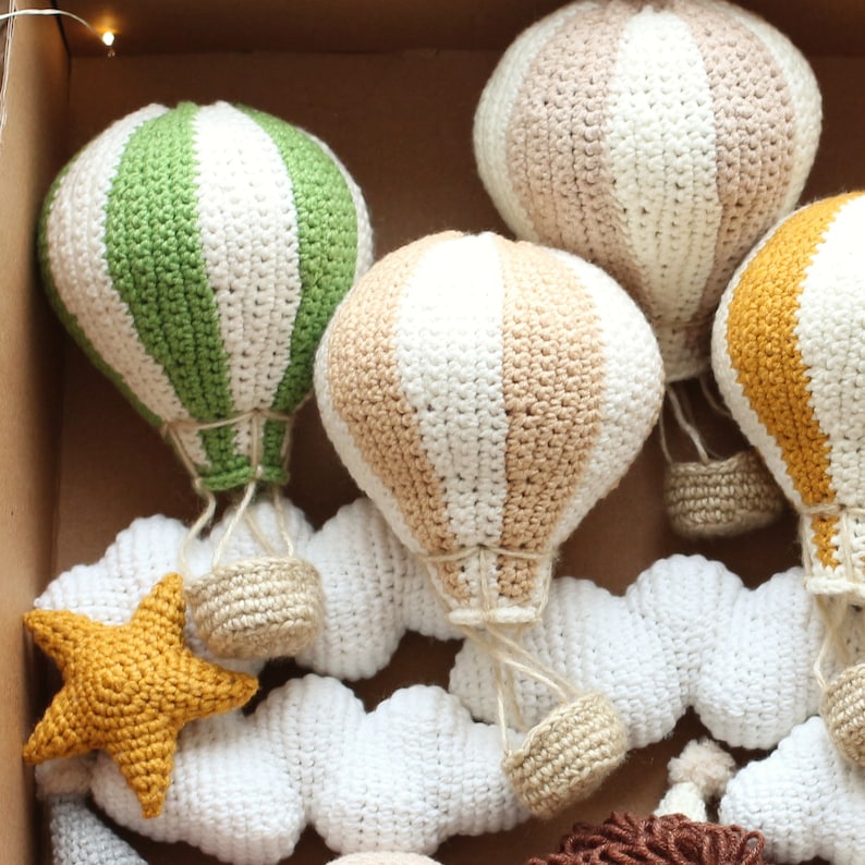 May include: A set of crocheted hot air balloons in green, beige, and yellow with white stripes, hanging from white crocheted clouds.
