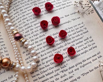 May include: A close-up of a book with a pearl necklace and red paper flowers. The book is open to a page with text about a pirate captain and a small island. The text reads, "Ah," said the captain, getting up. "D'you hear that, [69]"