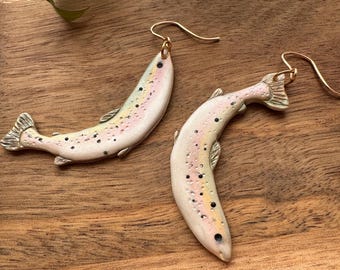 May include: A pair of dangle earrings featuring a realistic hand-painted trout design. The fish are painted in shades of pink, white, and brown with black spots. The earrings are made of polymer clay and have gold-toned hooks.