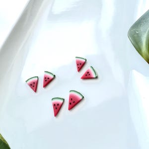 May include: Six miniature watermelon slice charms. Each charm has a red interior with black seed details, a white rind, and a green outer edge. The charms are arranged on a white surface.