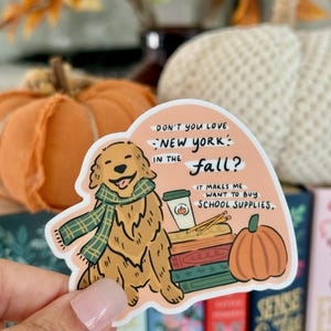 May include: A sticker featuring a cartoon golden retriever wearing a green scarf. The sticker has text that reads "Don't you love New York in the fall? It makes me want to buy school supplies." A pumpkin and coffee cup are also illustrated.
