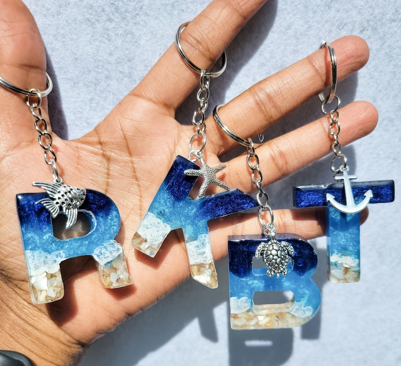 May include: Four resin letter keychains spelling out "RBT" and "T". Each letter features a blue and white ocean-themed design with silver charms of a fish, starfish, turtle, and anchor. The keychains are attached to silver keyrings.