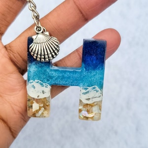 May include: A keychain featuring the letter "H" in resin, with a beach-themed design. The top of the letter is a deep blue, transitioning to a lighter blue and white, with sand and shells at the base. A silver seashell charm hangs from the chain.