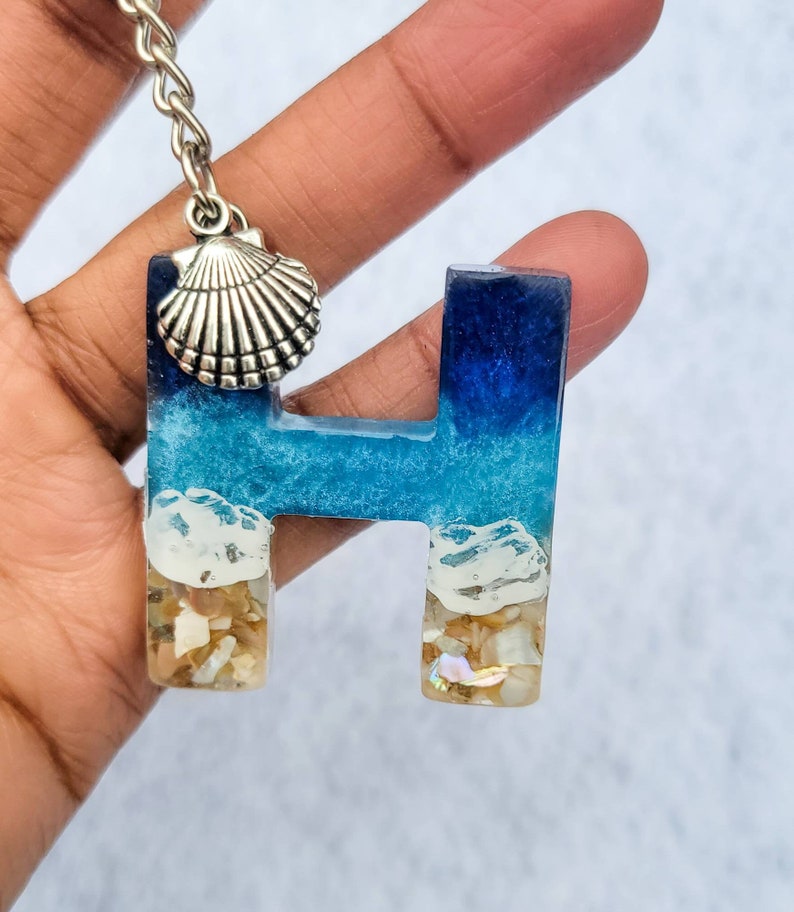 May include: A keychain featuring the letter "H" in resin, with a beach-themed design. The top of the letter is a deep blue, transitioning to a lighter blue and white, with sand and shells at the base. A silver seashell charm hangs from the chain.