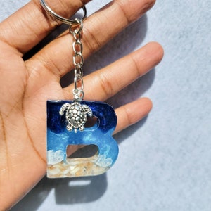 May include: A resin keychain in the shape of the letter "B". The keychain features a beach scene with blue and white resin representing the ocean, sand, and a silver turtle charm. The keychain is attached to a silver keyring.