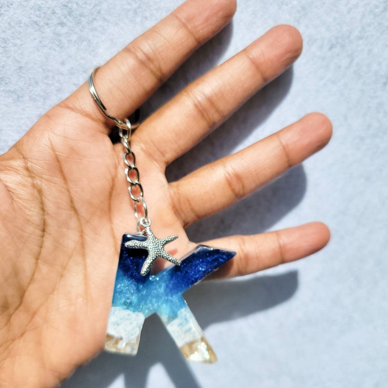 May include: A resin keychain in the shape of the letter "K" with a beach theme. The keychain features layers of blue, white, and tan resin, with a silver starfish charm. The keychain is attached to a silver chain and keyring.