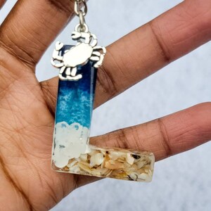 May include: A resin keychain in the shape of the letter "L" with a beach theme. The top portion is blue and white, resembling ocean waves, and the bottom is filled with seashells. A silver crab charm hangs from a chain.