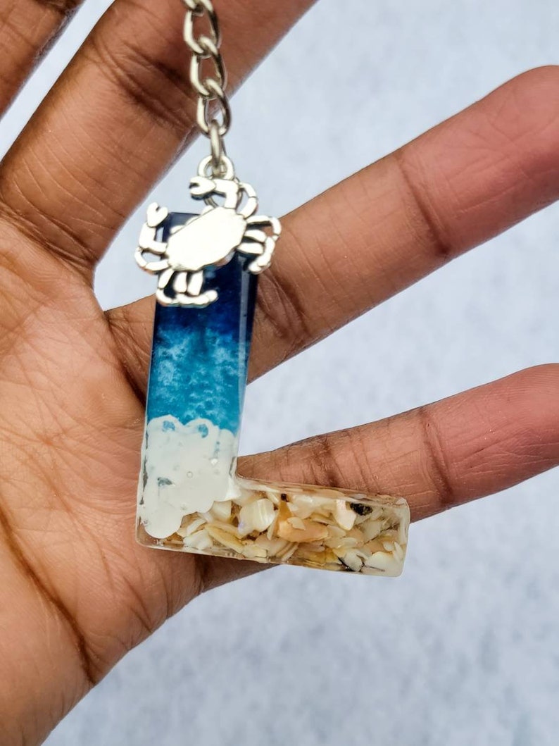 May include: A resin keychain in the shape of the letter "L" with a beach theme. The top portion is blue and white, resembling ocean waves, and the bottom is filled with seashells. A silver crab charm hangs from a chain.