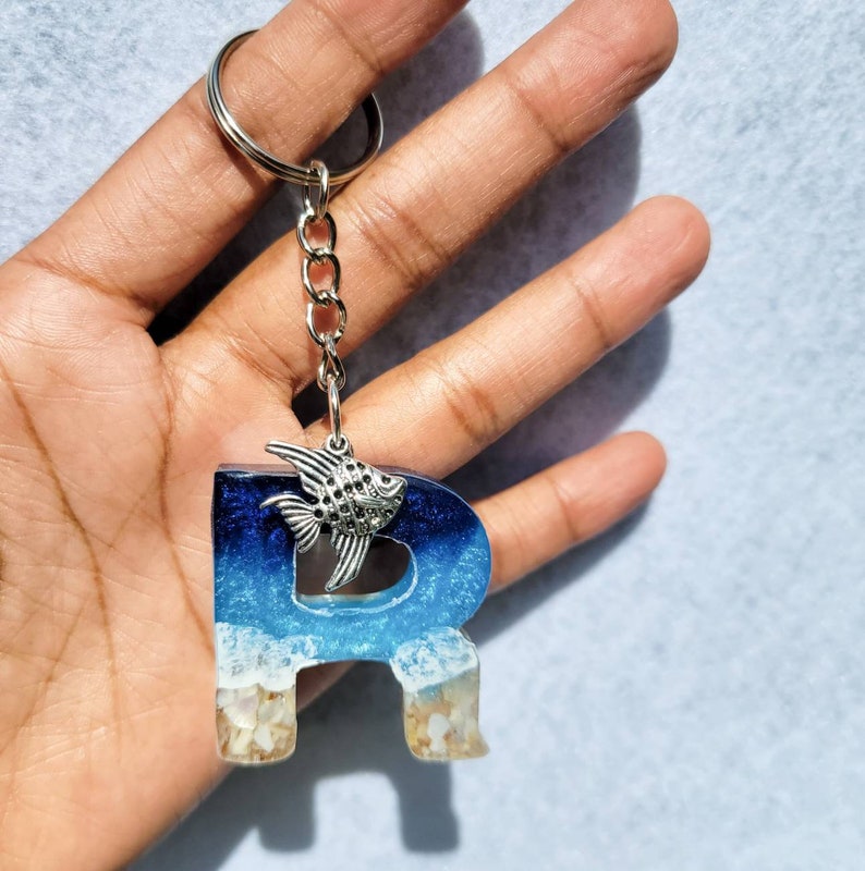 May include: A resin keychain in the shape of the letter "R" with a beach theme. The letter features blue and white resin, with a silver fish charm and a silver keyring. The bottom of the letter has a sand-like appearance.