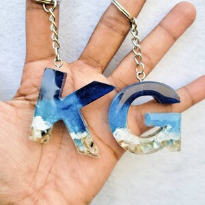 May include: Two resin keychains, featuring the letters "K" and "G". The letters are filled with blue and white resin, resembling an ocean scene with sand and shells at the base. Each keychain has a silver chain and ring.
