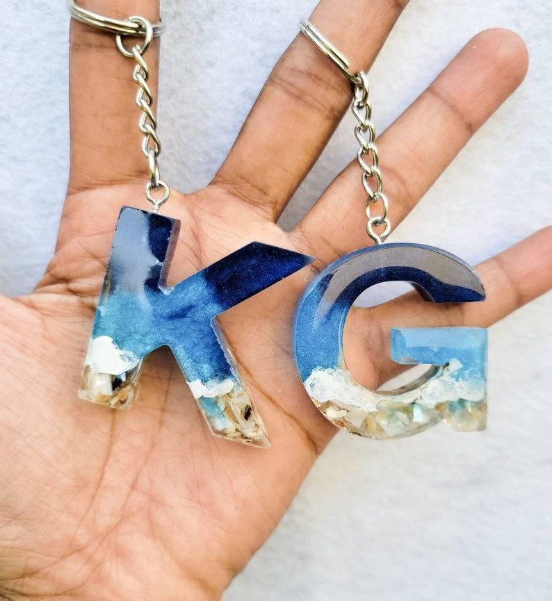 May include: Two resin keychains, featuring the letters "K" and "G". The letters are filled with blue and white resin, resembling an ocean scene with sand and shells at the base. Each keychain has a silver chain and ring.