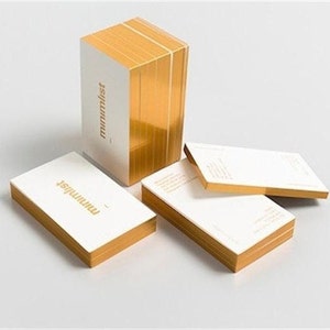 May include: A stack of white business cards with gold edges and the word "minimalist" printed in gold. Several cards are fanned out, showcasing the gold-colored edges. The cards are arranged on a white surface.