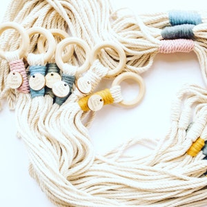 Può includere: Un set di cinque portavasi in macramè con anelli in legno. Ogni portavasi ha un fiocco di colore diverso con un'etichetta in legno su cui è scritto "Oak Tree Thread".