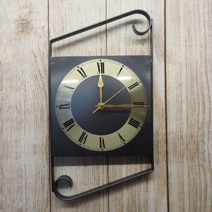 May include: A black square clock with Roman numeral markings and gold hands, mounted on a black metal frame with decorative scrolls. The clock is hanging on a light wood wall.