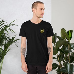 Puede incluir: Una camiseta negra con un contorno dorado de una pila de tres vasos en la parte delantera.