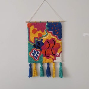 Puede incluir: Un tapiz de pared abstracto colorido con una variedad de formas y colores, incluyendo amarillo, naranja, rosa, azul y verde. El tapiz tiene una barra de madera en la parte superior y borlas en la parte inferior.