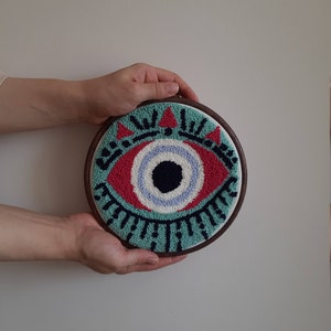 May include: A hand-crafted wall hanging featuring a colorful eye design with red, blue, white, and black yarn. The eye is surrounded by a teal and black pattern. The design is mounted in a wooden hoop.