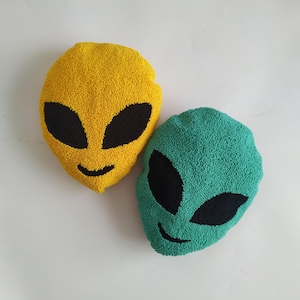 May include: Two plush alien-shaped cushions, one yellow and one green, with black embroidered eyes and a smiling mouth.