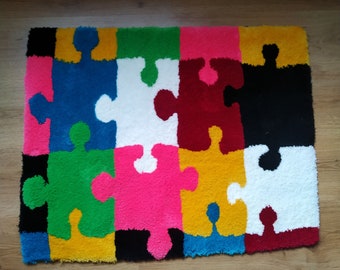 May include: A colourful rug made of yarn with a jigsaw puzzle piece design. The rug is made up of 12 jigsaw pieces in different colours, including blue, green, yellow, red, pink, white, and black.