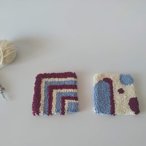 May include: Two small square rug samples with different patterns. One rug is white with blue and burgundy stripes, the other is white with blue and burgundy abstract shapes. A ball of white yarn and a rug hooking tool are also pictured.