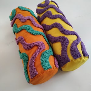 May include: Two cylindrical decorative pillows. One is orange with teal and purple wavy patterns. The other is yellow with purple wavy patterns. The pillows are made of fabric and have a textured, raised design.