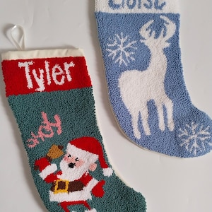 Può includere: Due calze di Natale. Una è verde acqua con un disegno di Babbo Natale e il nome "Tyler" in bianco. L'altra è blu con una renna bianca e il nome "Eloise". Entrambe hanno la parte superiore bianca e anelli per appenderle.