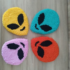 May include: Four colorful felt alien head coasters. The coasters are shaped like alien heads with black eyes and a black mouth. The coasters are yellow, blue, pink, and orange.