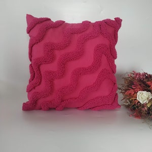 May include: A pink throw pillow with a textured wave pattern. The pillow is made of a soft, plush fabric.