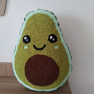 Puede incluir: Una almohada de felpa con forma de aguacate verde con un hueso marrón y una cara sonriente. La almohada tiene un contorno verde claro.