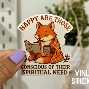 May include: A vinyl sticker featuring a cartoon fox reading a book and holding a mug. The fox is wrapped in a plaid blanket. The sticker has the text "Happy are those conscious of their spiritual need."