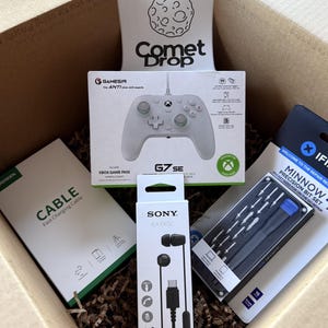 May include: A cardboard box containing a GameSir G7 SE controller box with a Comet Drop sticker, a UGREEN fast charging cable box, a Sony IER-EX15C earphone box, and an iFixit Minnow precision bit set in its retail packaging, all nestled in paper filler.