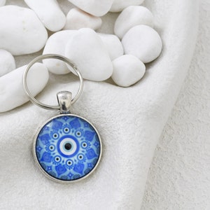 May include: A silver keyring with a circular pendant displaying a blue and white eye motif. The pendant showcases a detailed pattern with a central black dot, encircled by concentric patterns and floral designs. The keyring is set against a white backdrop.