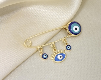 Puede incluir: Alfiler de seguridad dorado con amuletos de ojo turco azules y blancos. El alfiler presenta un ojo grande en la parte superior y tres colgantes con diseños de ojos. El alfiler está sobre una tela color crema.