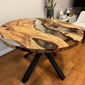 May include: Round wooden dining table with a clear epoxy resin top. The table has a natural wood grain pattern and black metal legs.