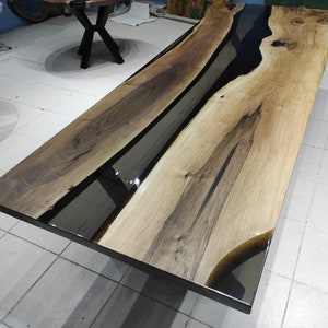 May include: A long wooden table with a black epoxy resin river running down the center. The table has a natural wood finish and is supported by black metal legs.