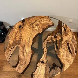 May include: A round wooden table top with a natural wood grain pattern and a clear epoxy resin fill. The table top is set on a black metal base.
