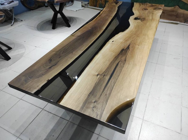 May include: A long wooden table with a black epoxy resin river running down the center. The table has a natural wood finish and is supported by black metal legs.