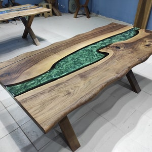 May include: A large wooden table with a live edge and a green epoxy river running through the center. The table has a natural wood finish and is supported by a black metal X-shaped base.