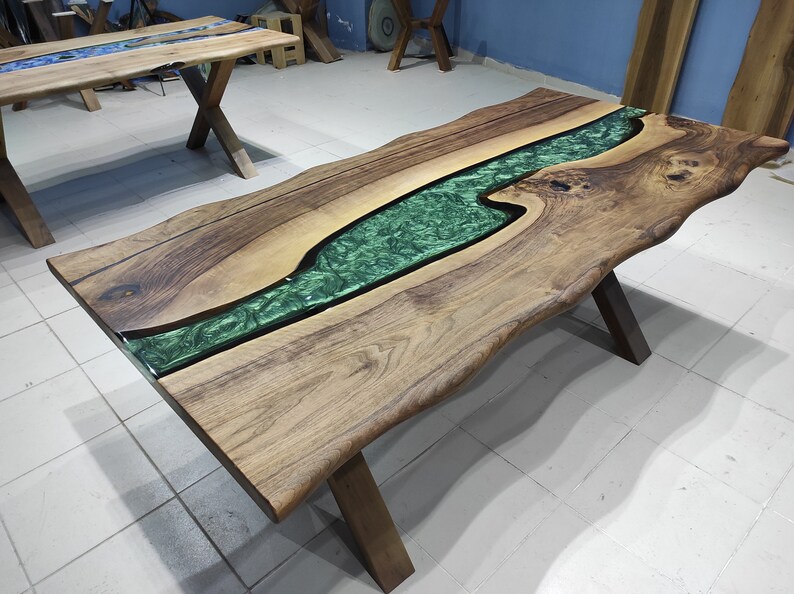 May include: A large wooden table with a live edge and a green epoxy river running through the center. The table has a natural wood finish and is supported by a black metal X-shaped base.