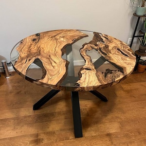 May include: Round wooden table with a clear epoxy resin top and a black metal base. The table top has a natural wood grain pattern.