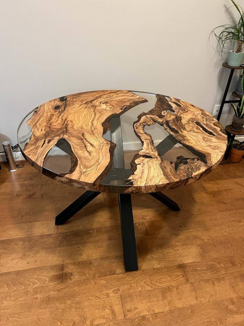 May include: Round wooden table with a clear epoxy resin top and a black metal base. The table top has a natural wood grain pattern.