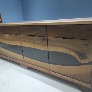 May include: A long, wooden sideboard with four drawers and three glass-fronted doors. The doors feature a unique design with a dark resin inlay that resembles a river flowing through the wood.