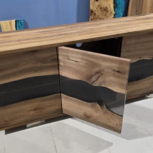 Puede incluir: Un mueble de televisión de madera con un acabado de vetas de madera natural y una incrustación de resina epoxi oscura. El mueble tiene dos puertas, una de las cuales está abierta, revelando un espacio interior. El mueble es moderno.