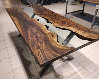Puede incluir: Una mesa de madera grande con un borde natural y un río de resina epoxi transparente que atraviesa el centro. La mesa está sostenida por una base en forma de X de metal negro.