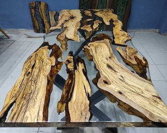 May include: A large, live edge wooden table with a clear epoxy resin fill. The wood is a light brown with dark brown streaks. The table has a natural, organic shape.