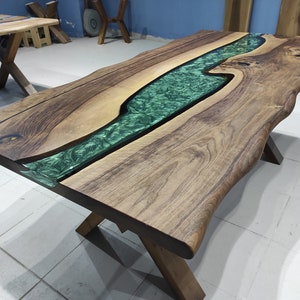 May include: A large wooden table with a live edge and a green epoxy river running through the center. The table is supported by a black metal X-shaped base.