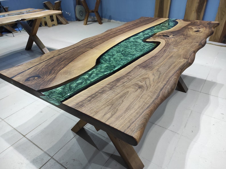 May include: A large wooden table with a live edge and a green epoxy river running through the center. The table is supported by a black metal X-shaped base.